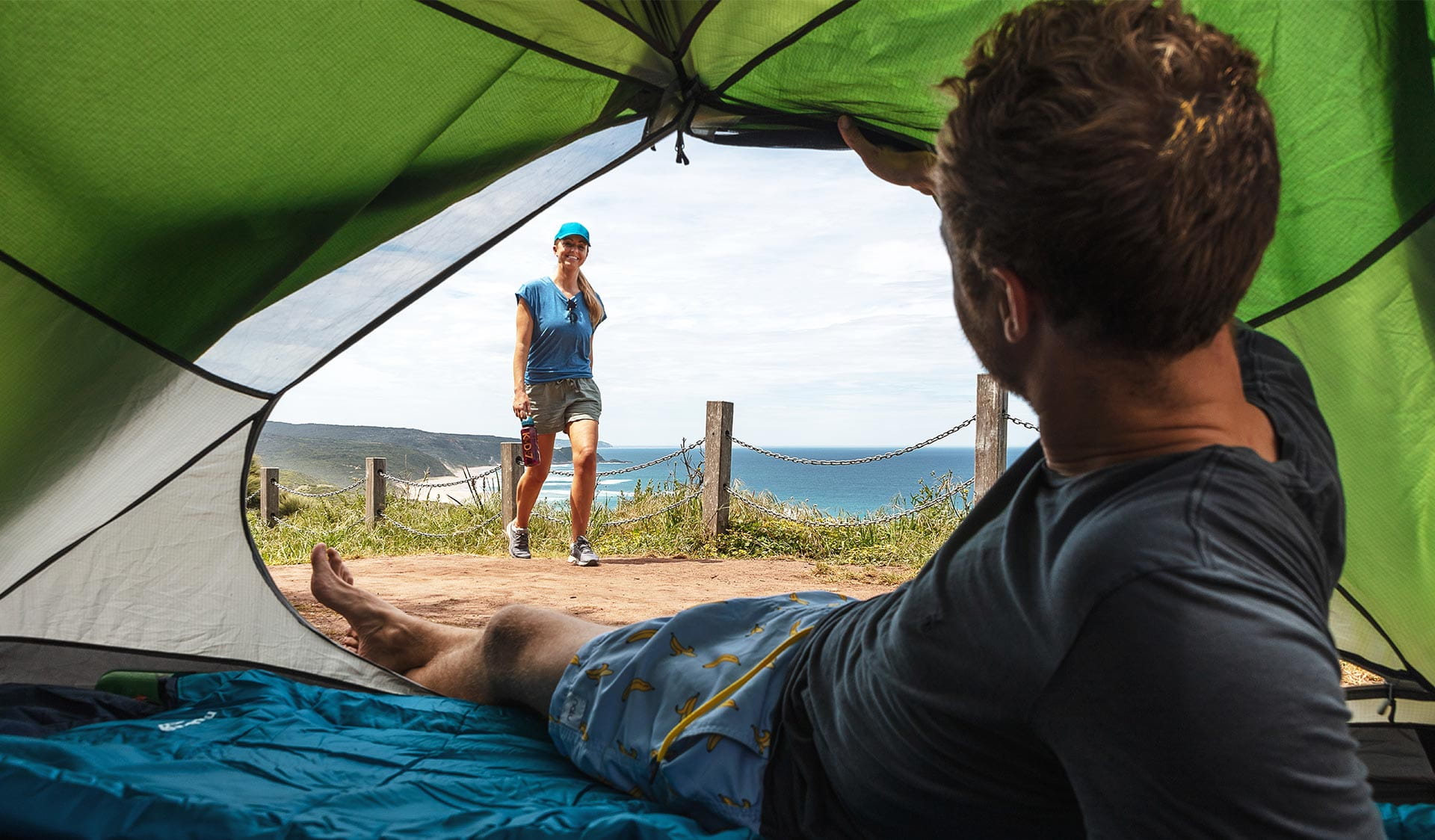 A man looks out of his tent towards his partner walking towards him with the coast in the background at Johanna Beach in the Great Otway National Park.