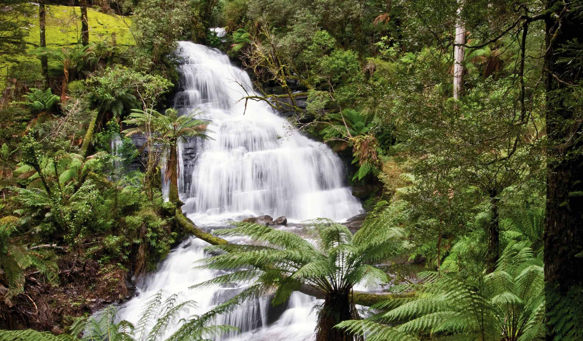 A young woman walks on rocks across the water in front of Triplet Falls.