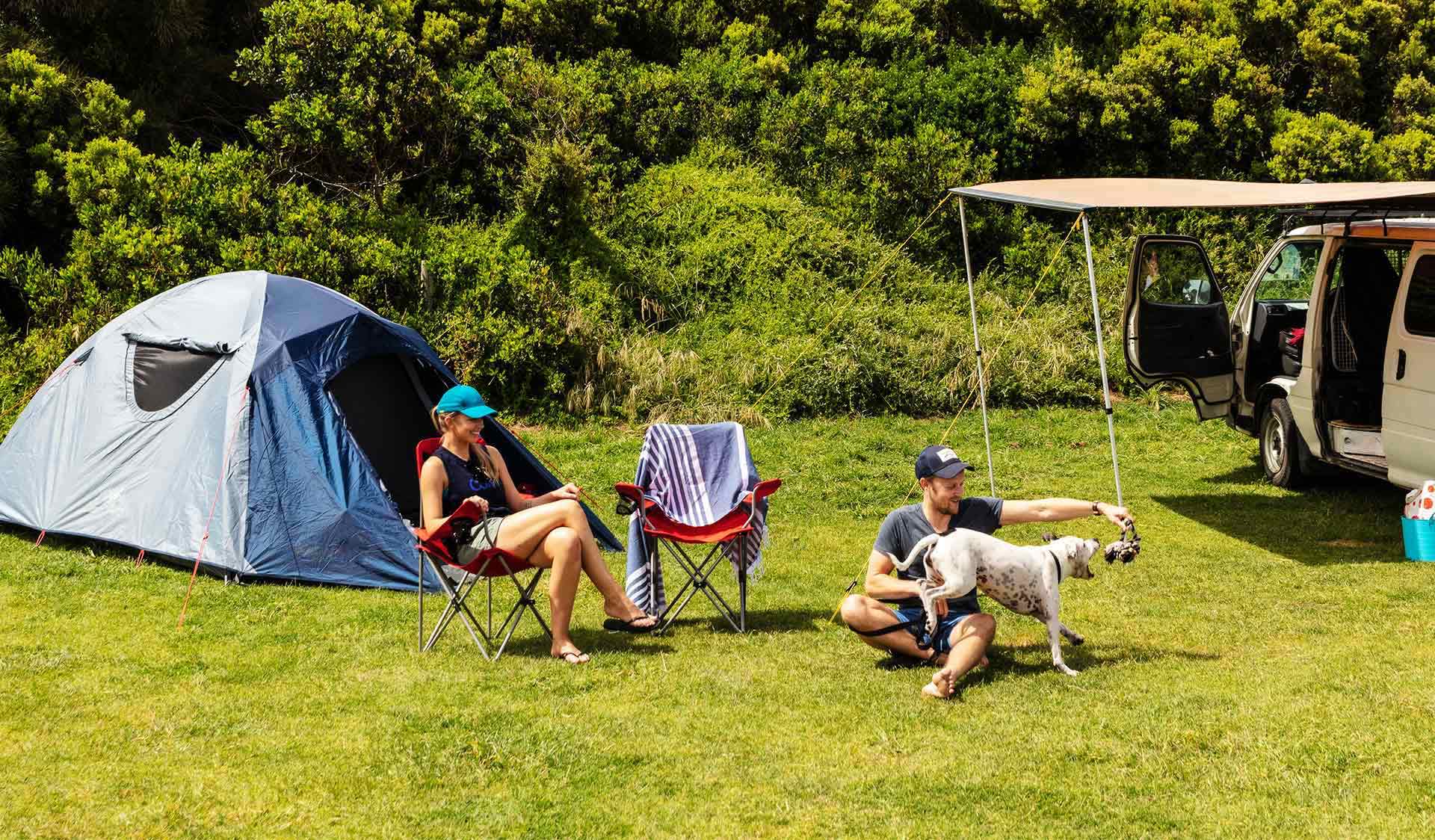 A couple in their thirties play with their dog at Johanna Beach Campground next to their tent and campervan.
