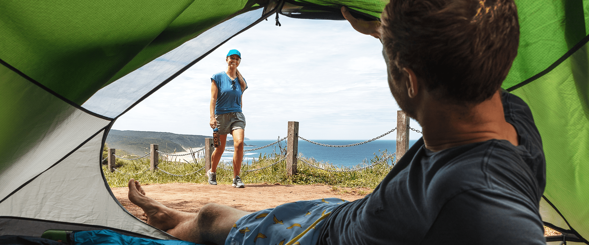 A man lays in his tent mattress looking out towards the ocean, his partner is walking towards the tent. 