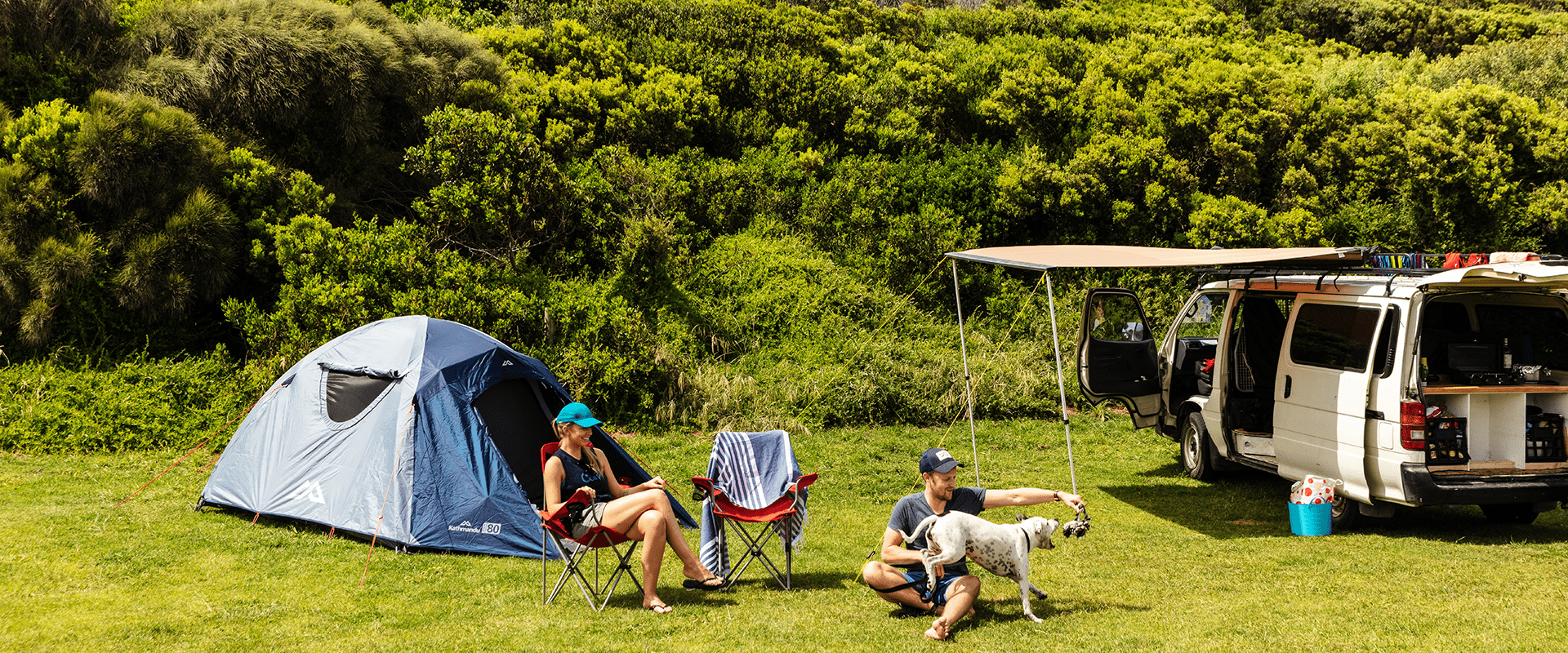 A couple sits around their tent playing with their dog, a white van is parked next to their tent, coastal vegetation surrounds them.