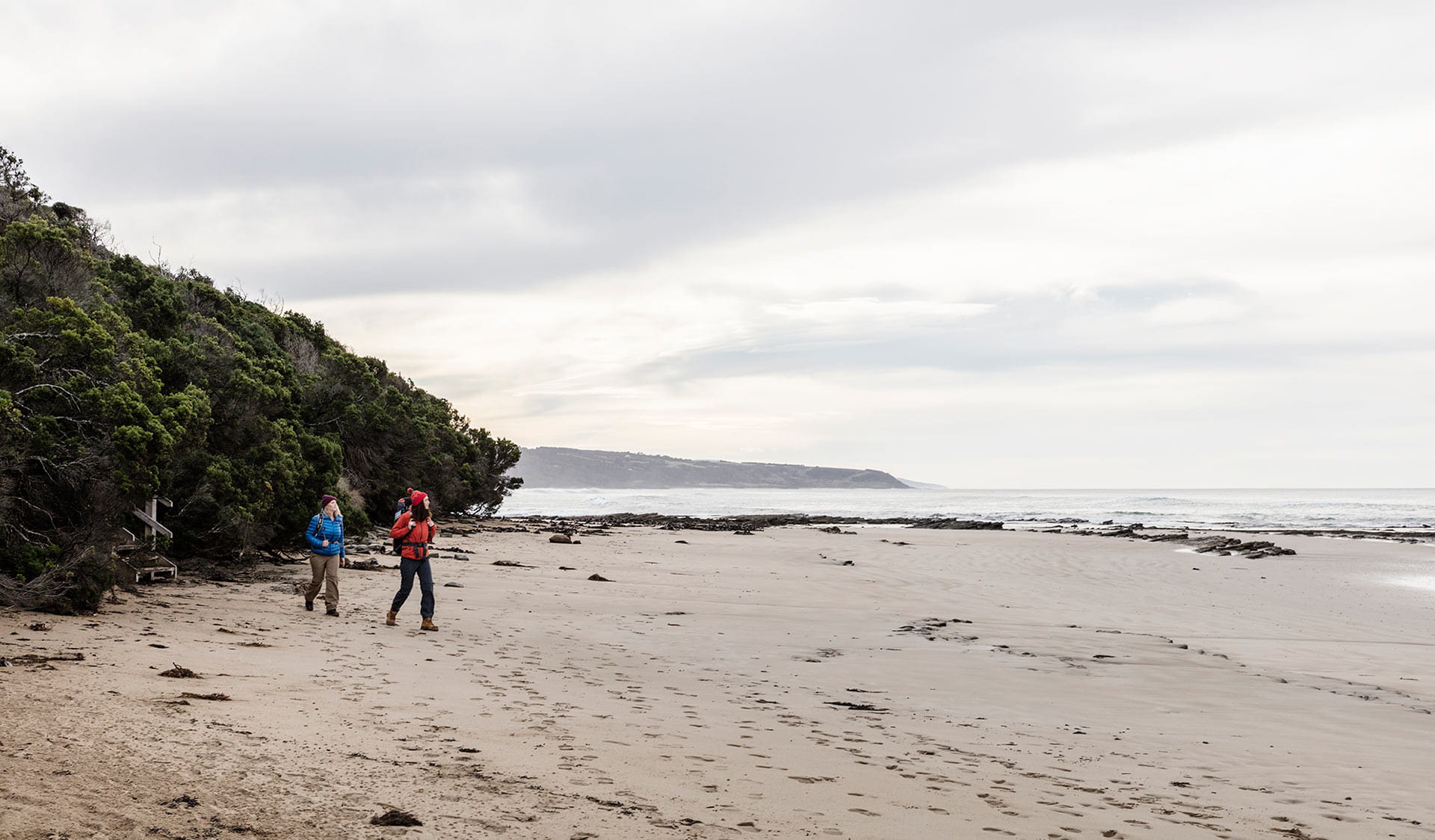 Two women walking the Great Ocean Walk step out of the Ti-Tree scrubb and onto the beach at Blanket Bay.