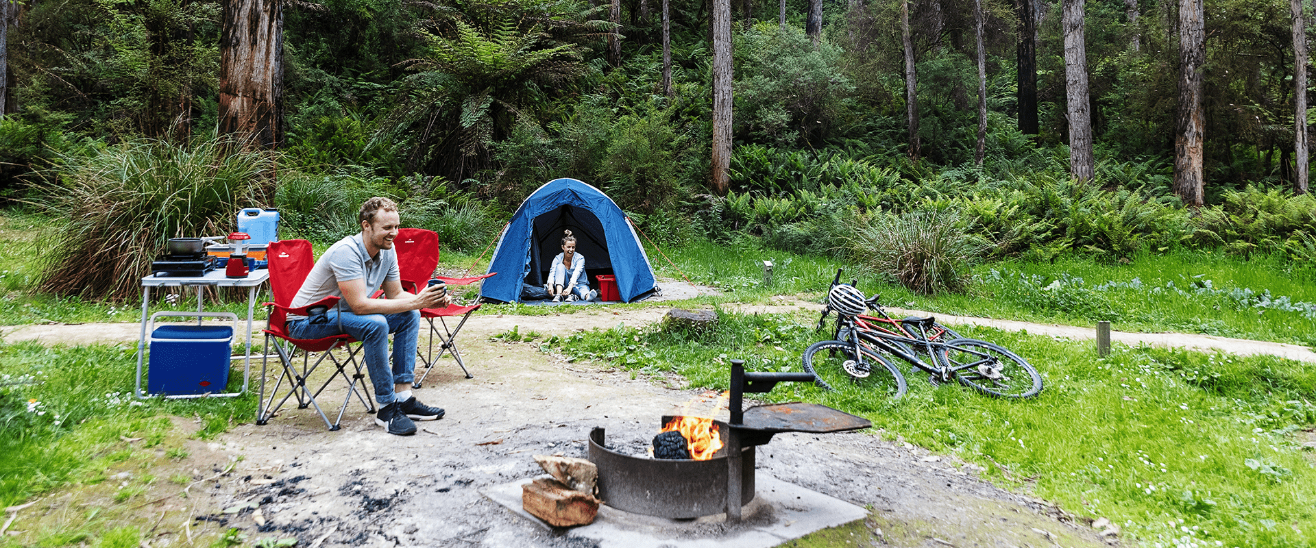 A man sits on a camping chair and a female peaks out from a dome camping tent, lush tree ferns and tall Eucalyptus dominate the background.