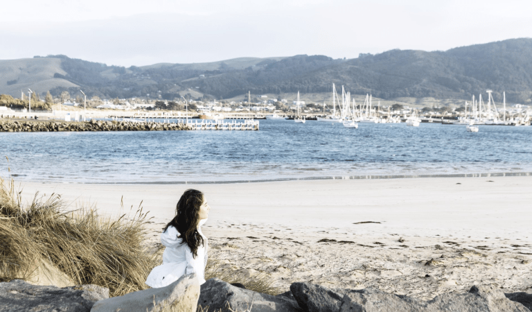 A woman sits on the beach, behind her is boats on the ocean and hills in the distance