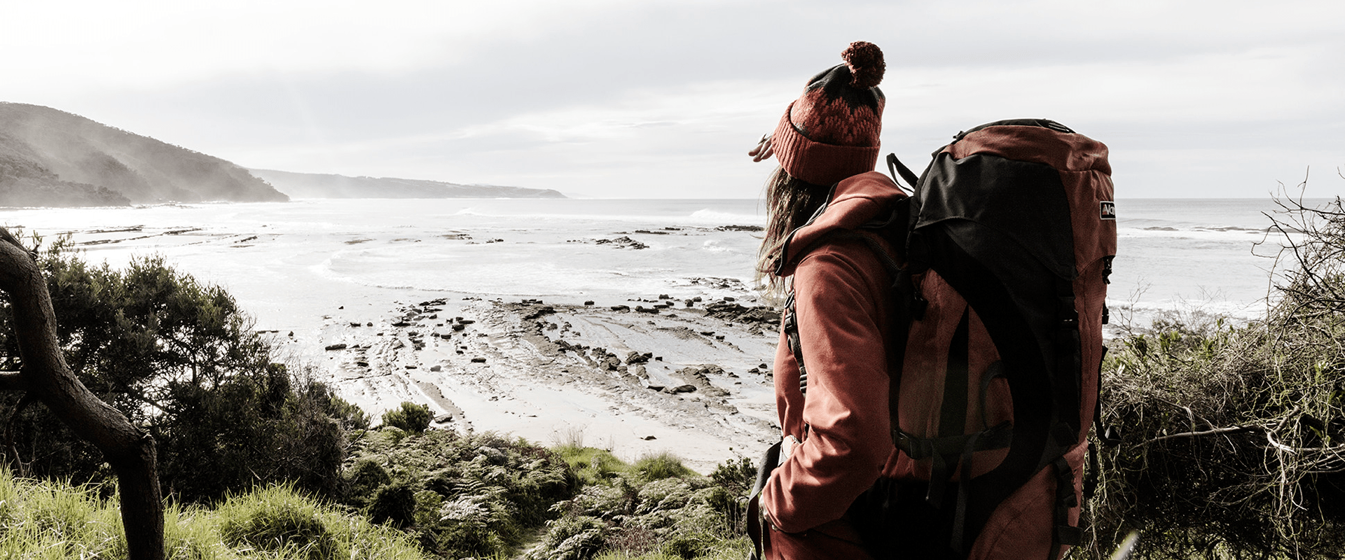 A hiker carrying an overnight pack looks forward to an ocean vista with a sandy path leading down to the beach.