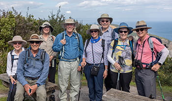 Group of people wearing backpacks, hats, and sunglasses posing together and smiling at the camera with the ocean view in the background.