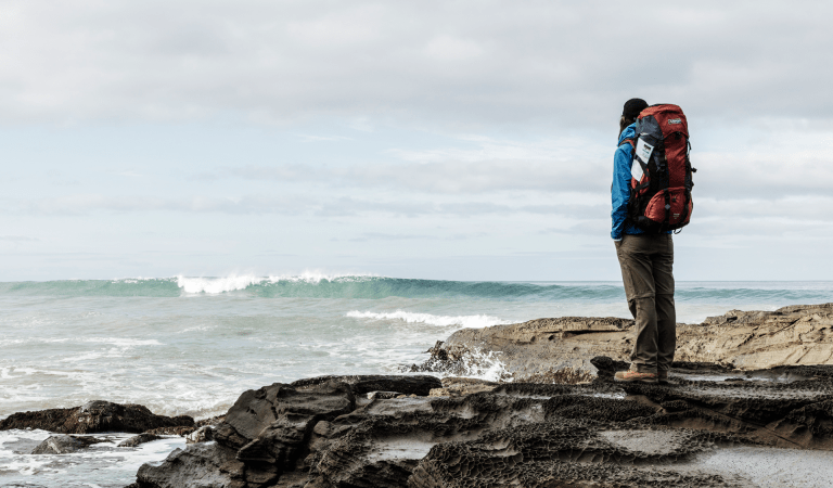 A woman with a large hike pack stands on a rock next to the ocean