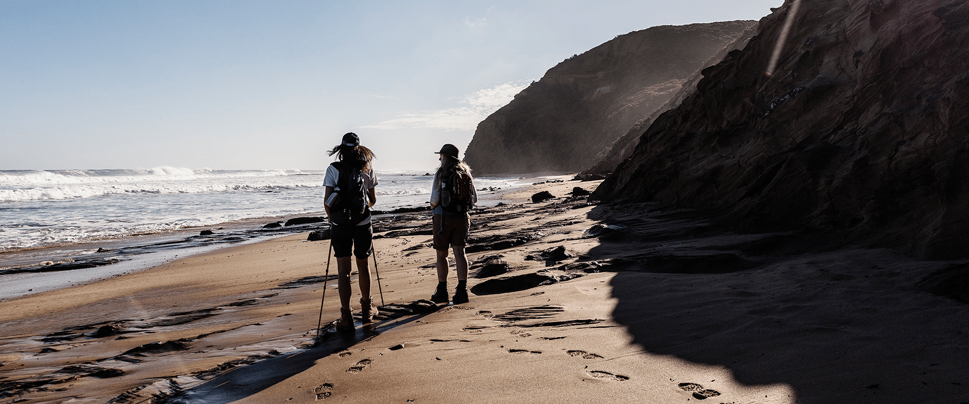 A couple carrying hiking backpacks and walking sticks walks on a sandy beach with waves on the left side and a rocky cliff on the right side.