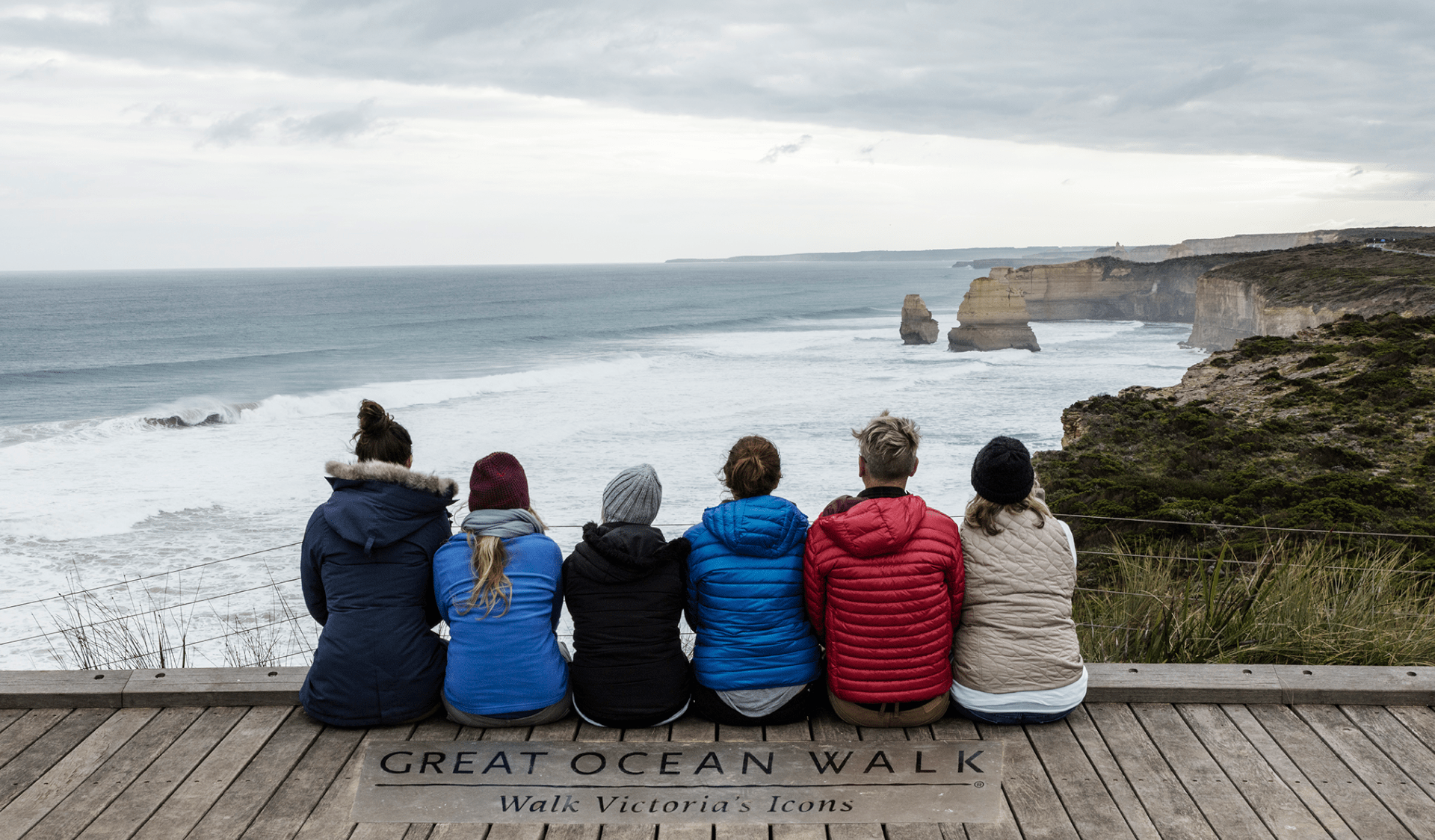 Six friends sit on a boardwalk that has Great Ocean Walk written on it. Their backs are to the camera and the Twelve Apostles are in front of them