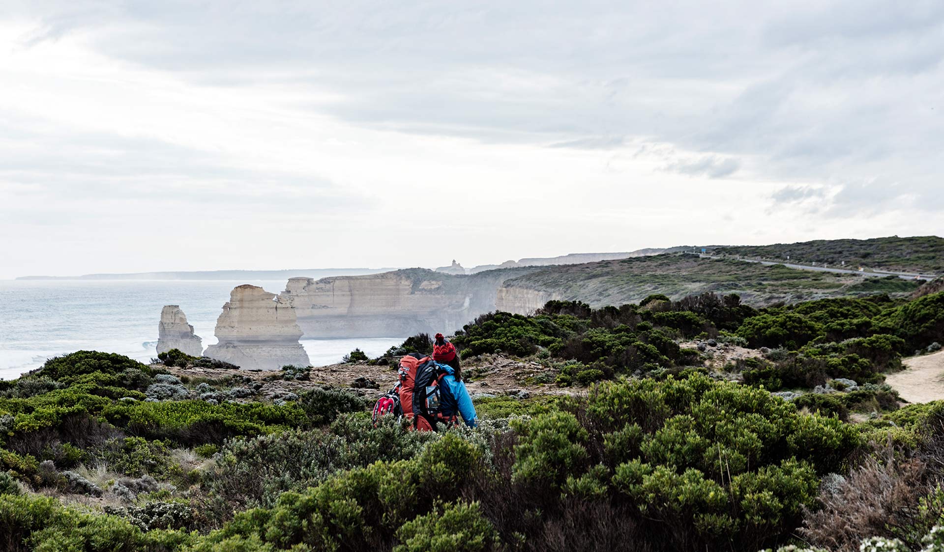 A women in a blue jacket walks through the heath on the cliffs above the Twelve Apostles.