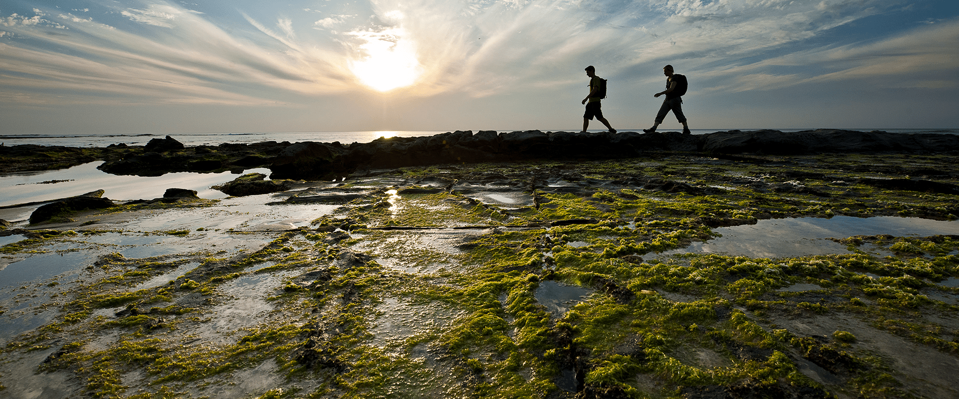 Two hikers walk across a rocky beach platform with a low sun behind them.