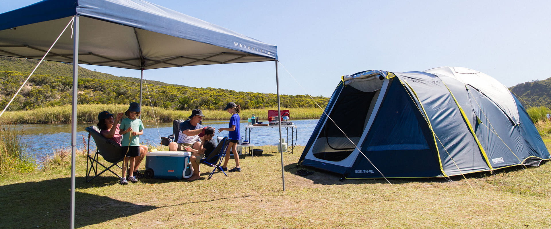 A family with three young children relax under a marquee shelter next to a river and alongside their blue family-sized tent.