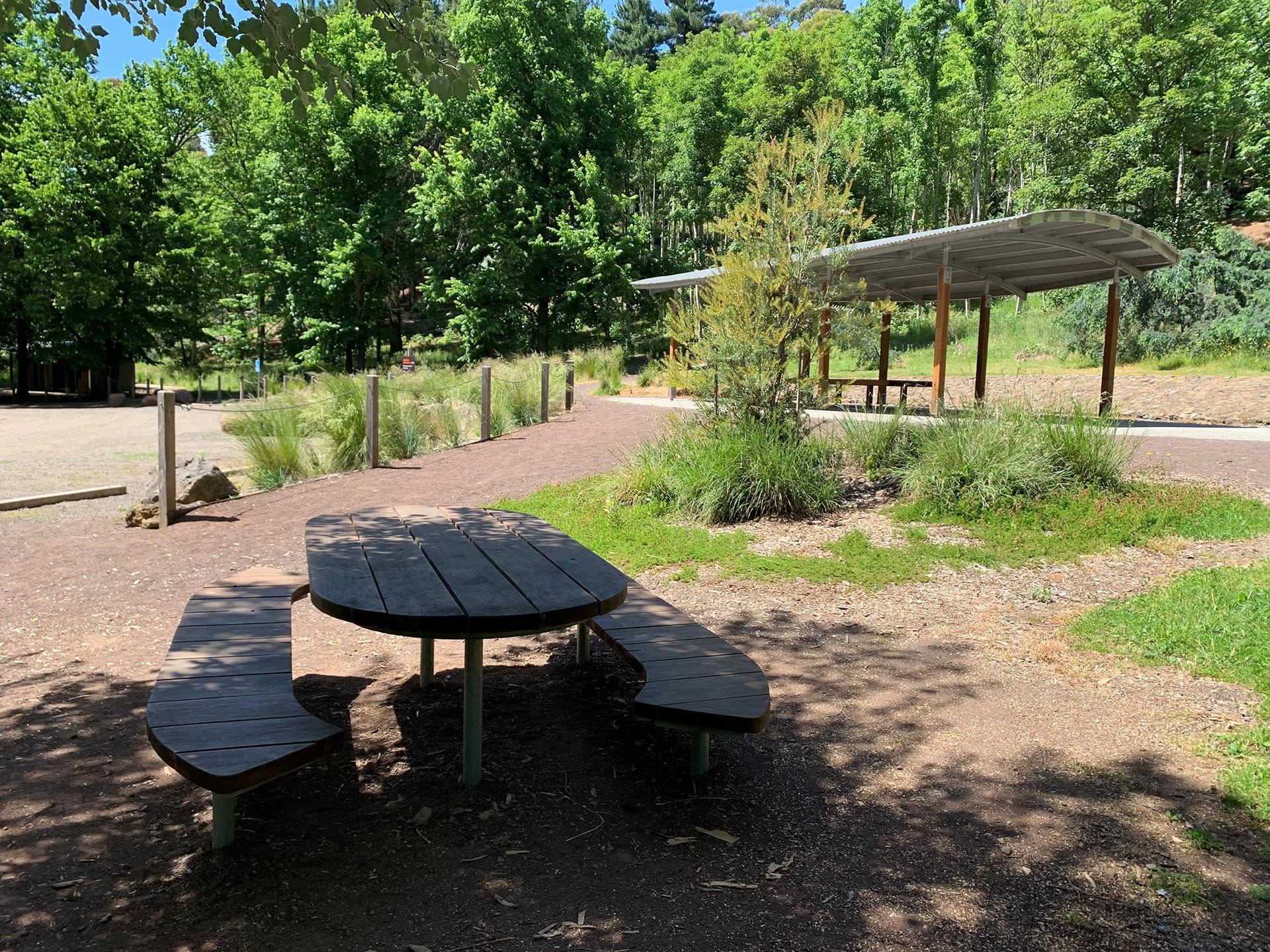 Wooden picnic table and benches with a shelter and trees in the background.
