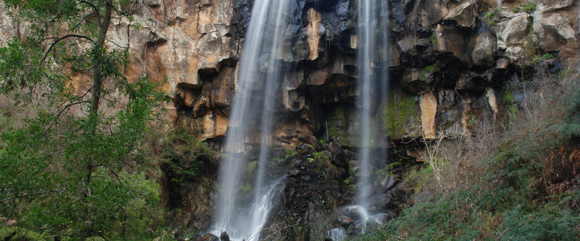 A full flowing waterfall cascades down into a rocky cliff face surrounded by rugged bushland.