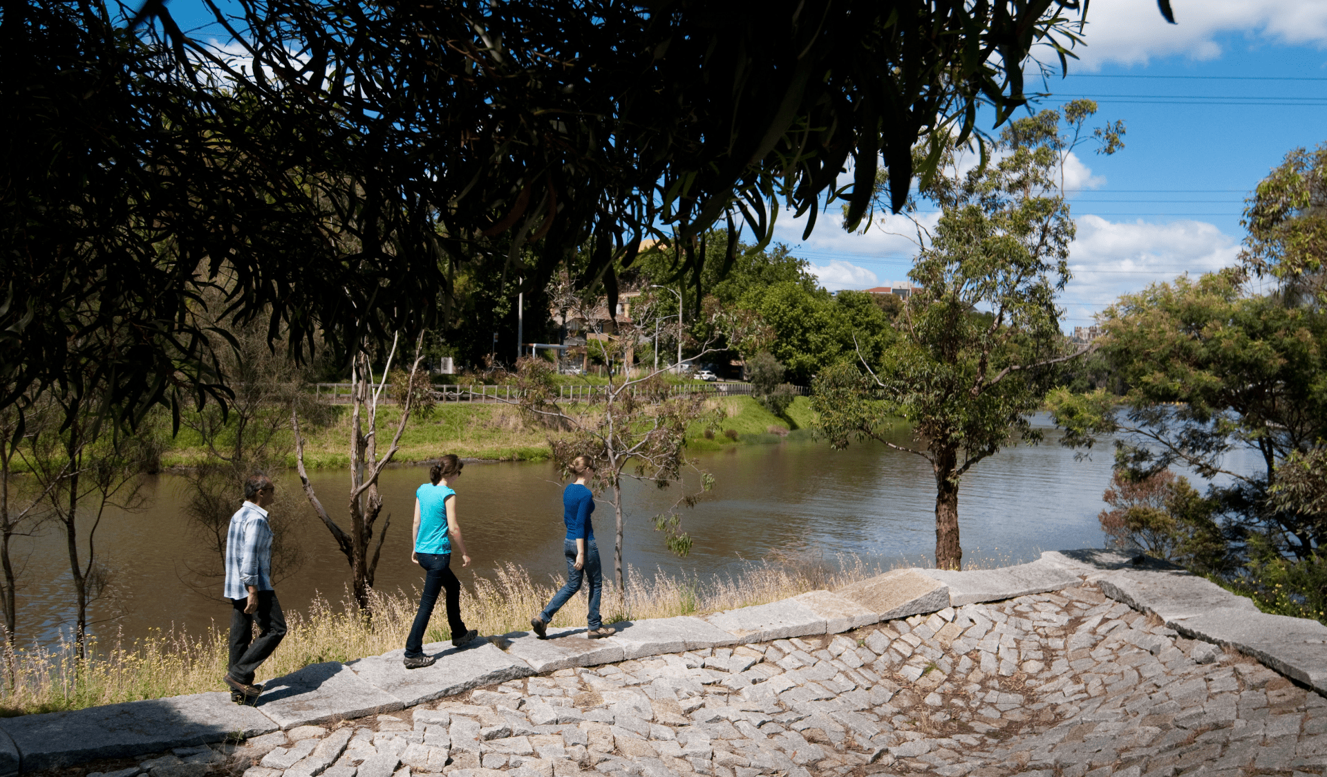 Three adults walk on a path alongside the Yarra River in Herring Island Environmental Sculpture Park