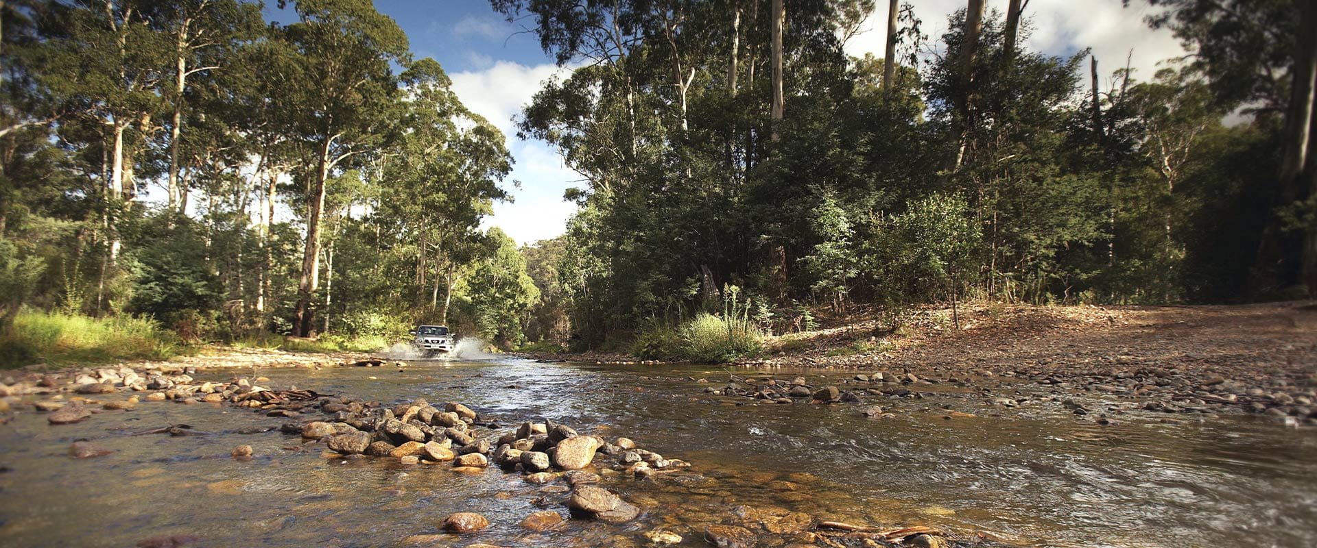 A silver 4WD crosses a shallow river, lined will small round rocks infront of backdrop of gum trees