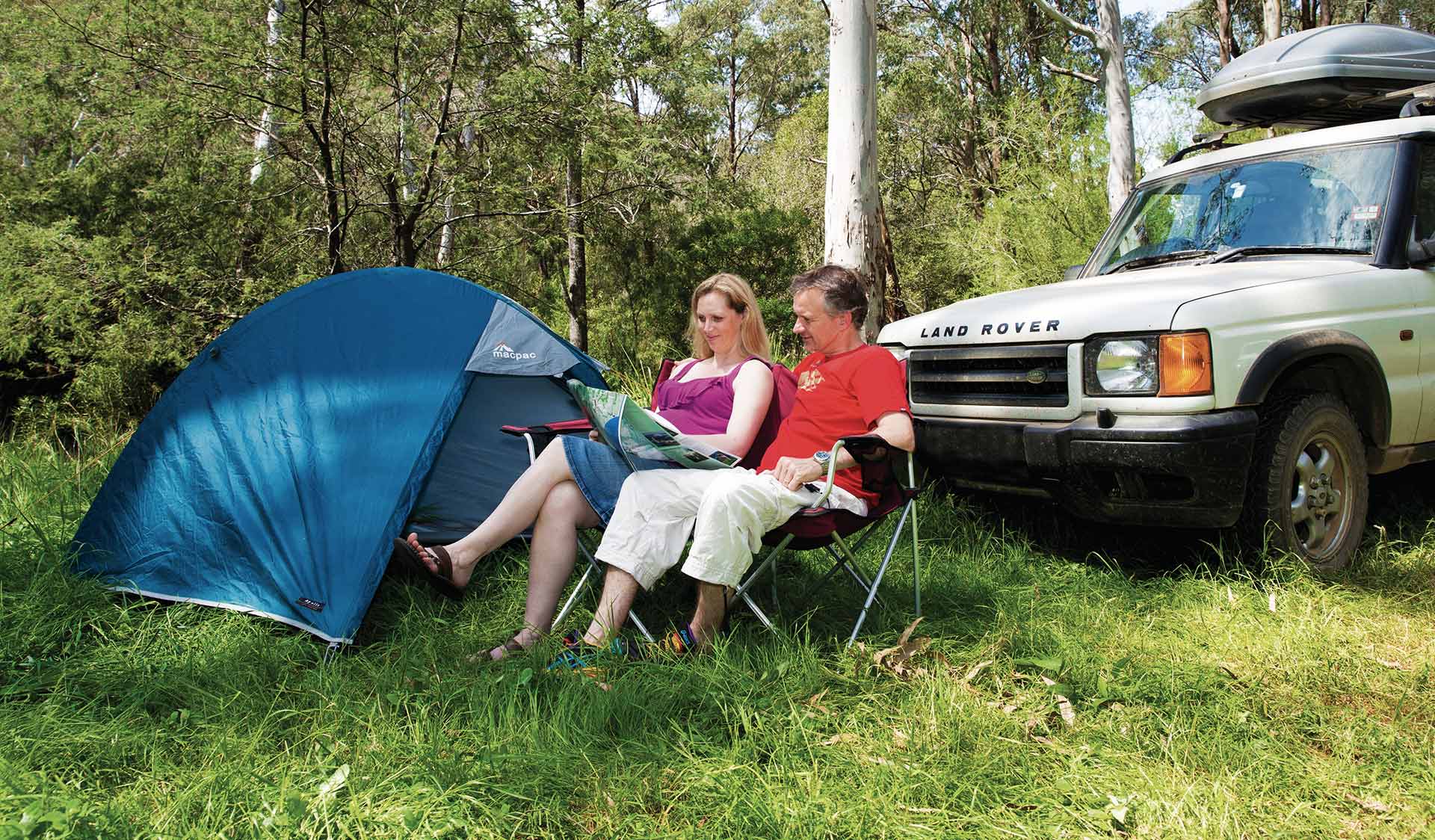 A couple sit and look at a map next to their tent and car.