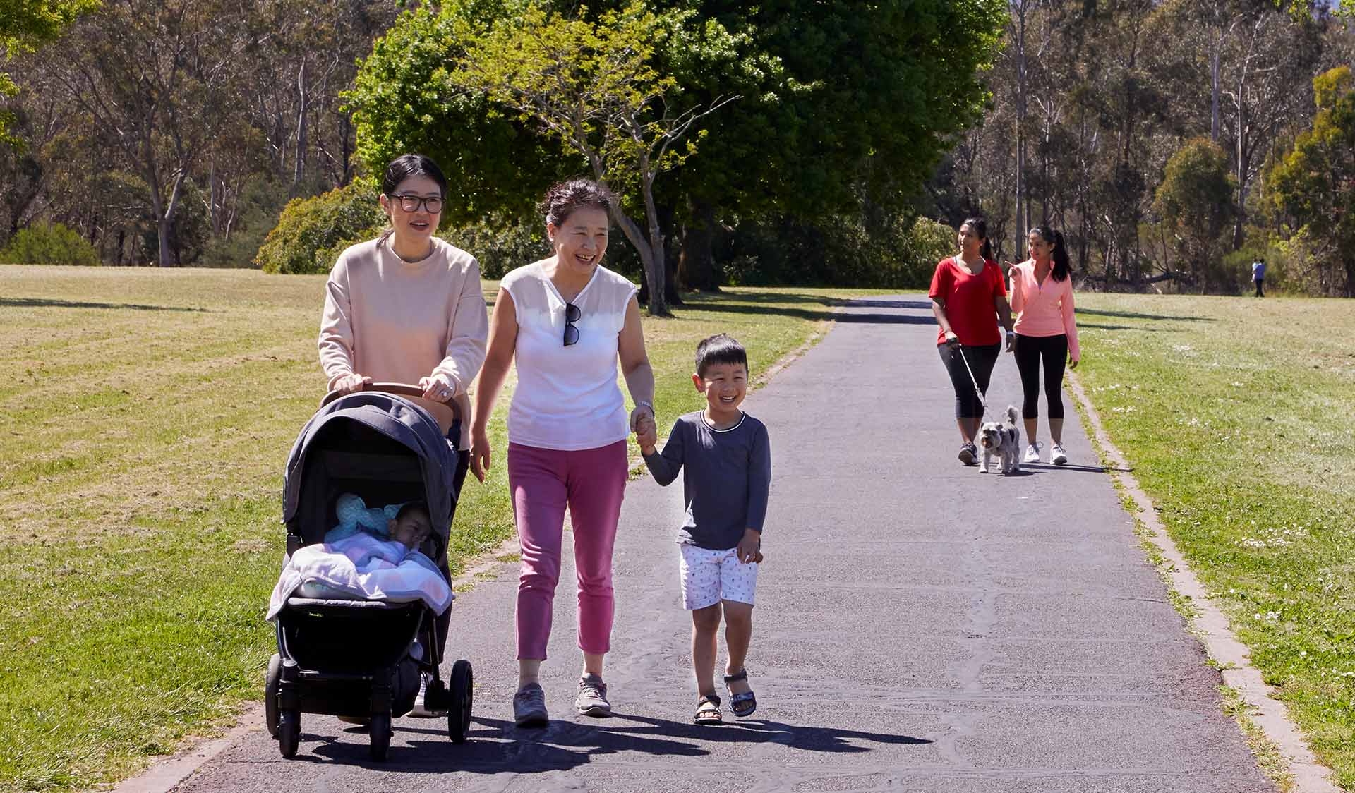 A young boy walks hand-in-hand with his grandmother, while his mother pushes his younger sibling in a pram. 