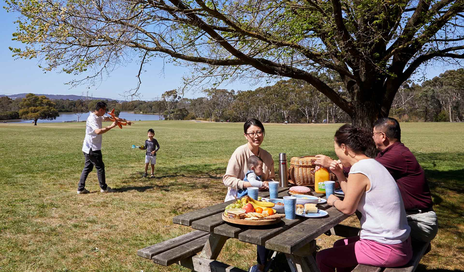 A family share a picnic while a father and son play with a model aircraft in the background.