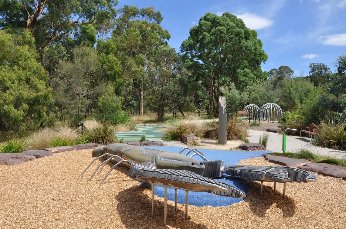 The giant yabby located at Jells Park Playscape. It is grey in colour and sits on top of a large tanbark pit. Bushland and other play structures are in the background.