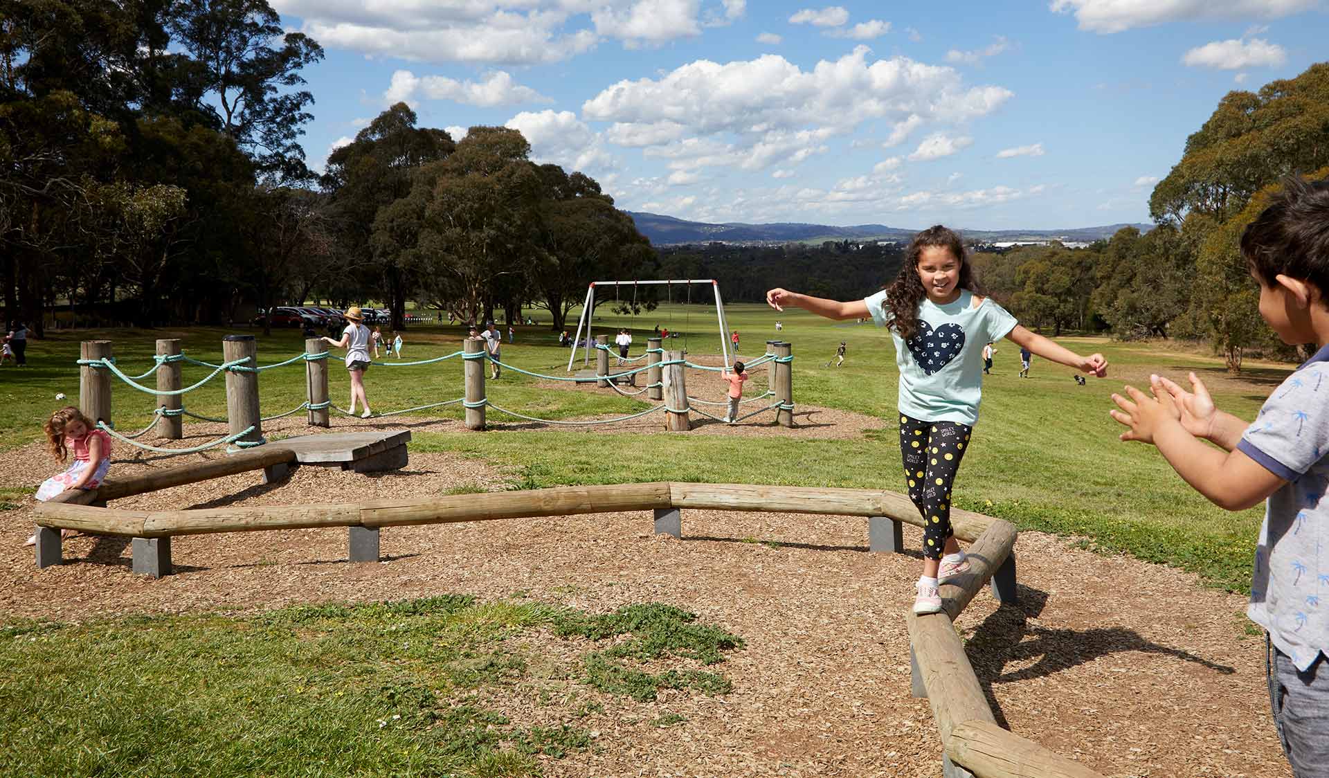 A young girl walking on a winding balance beam with children and families playing in the background at Yabby Hill Playscape