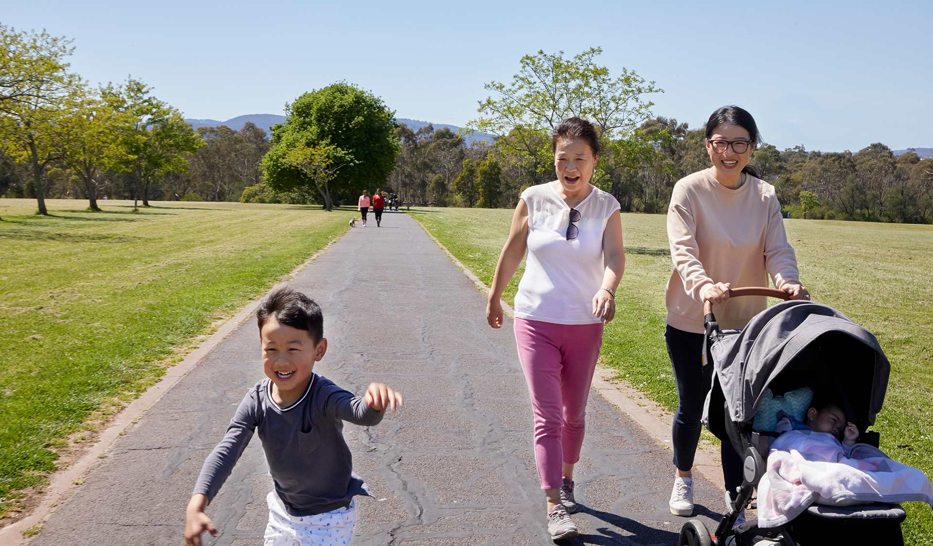 A grandmother watching her grandson happily walking while his mother pushes his younger brother in a pram at Jells Park