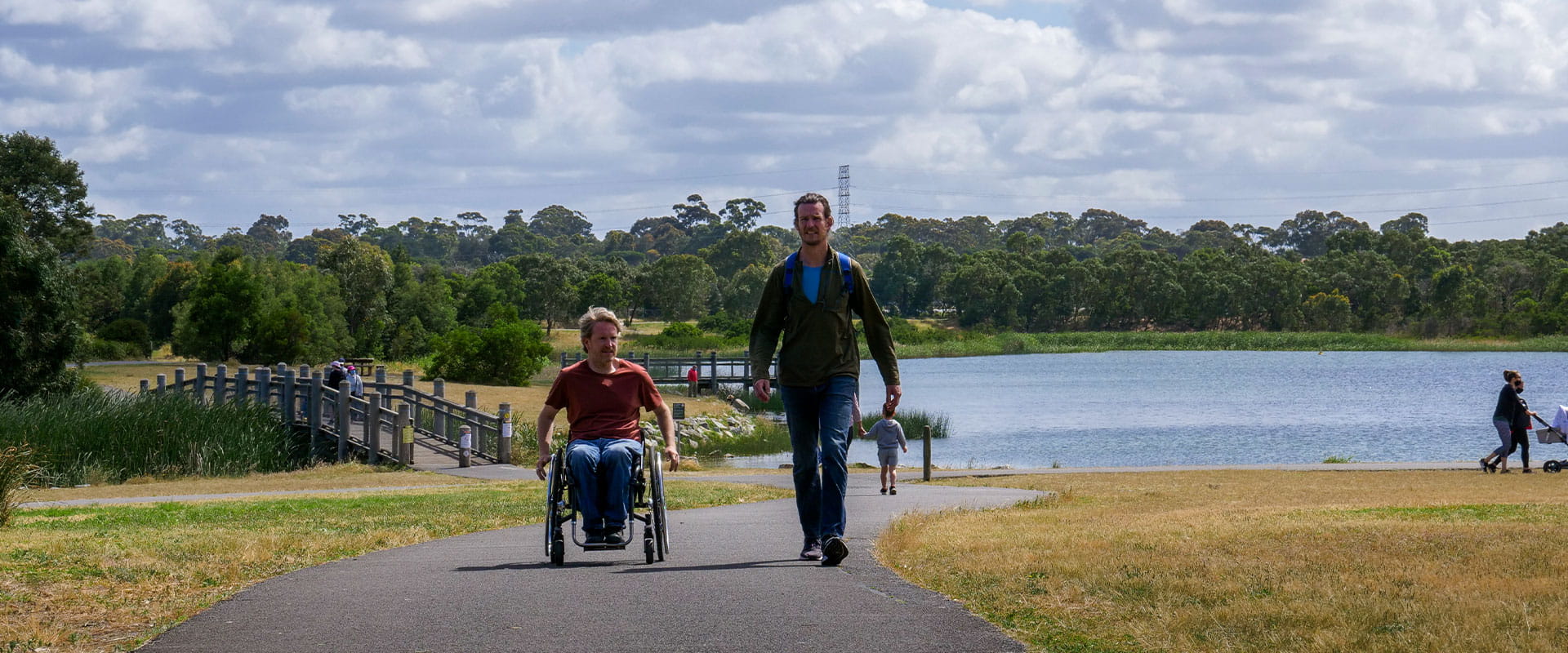 Two men, one of whom is in a wheel chair, walk up a sealed path, away from a lake in the background.