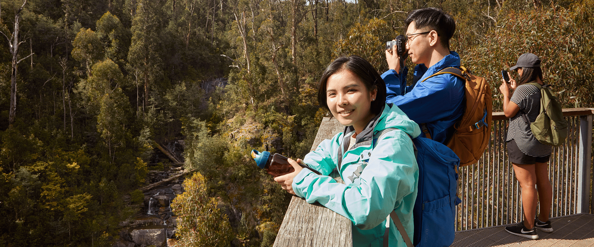 A group of people standing on a wooden viewing platform looking out towards a large waterfall surrounded by tall Eucalyptus trees. A lady smiles at the camera while her partner takes a photo of the scene. 
