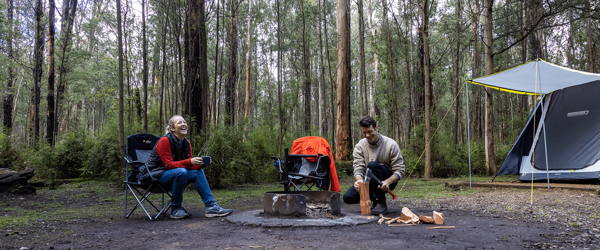 Two campers sit around a campfire, the male is splitting kindling to start a campfire. A tent is surrounded by tall trees and bush.