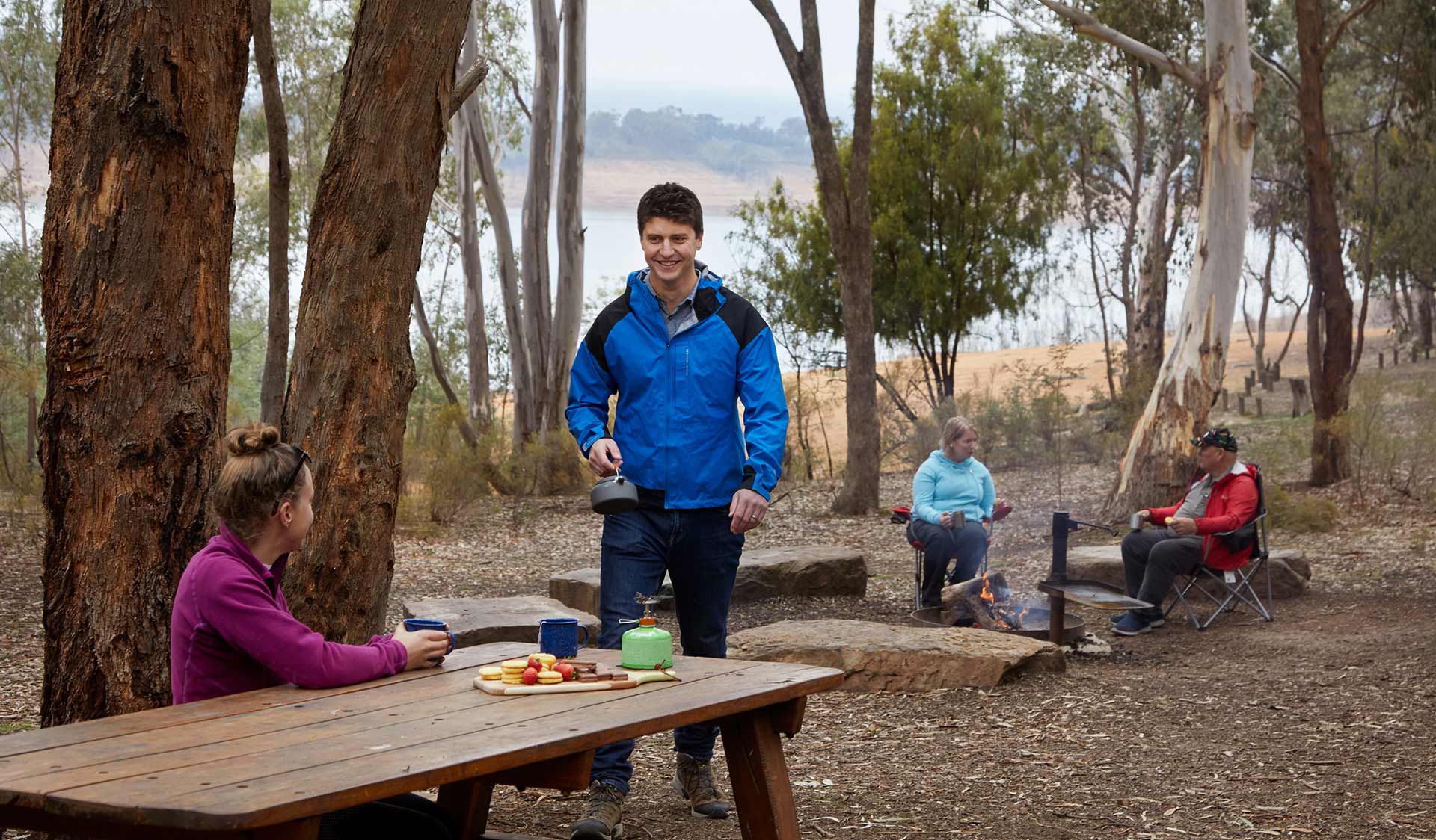 A man brings a kettle to the picnic table where his partner is sitting, while an older couple sit around a campfire in the background.