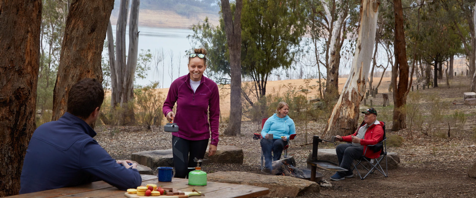 A family gather around a picnic table in a peaceful forest landscape. A lake in the background. 