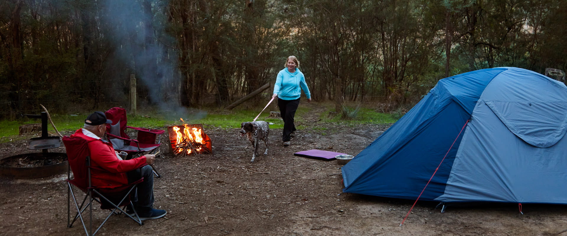Two people camping in a bushland landscape. One walks a dog and the other sits by the camp fire. A tent nearby.