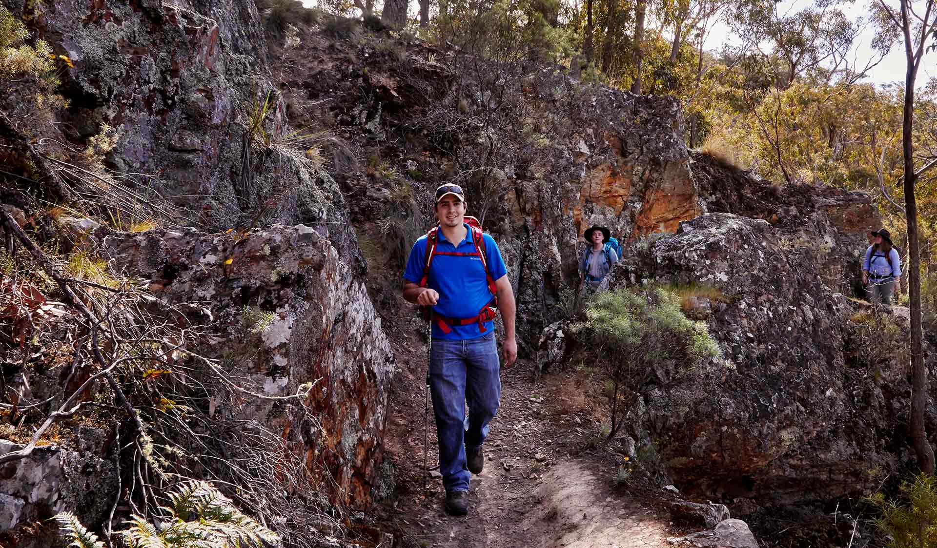 A man wearing a blue shirt and jeans walks along the Back Byers Track with two companions.