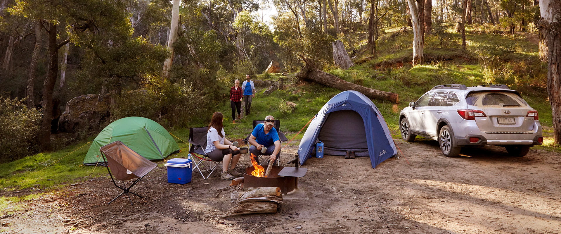 A man and woman holding cups and a bowl sit on camp chairs by a lit fire pit between two tents and a car. The man feeds the fire as the woman turns to another man and woman walking towards them along a grass track. 