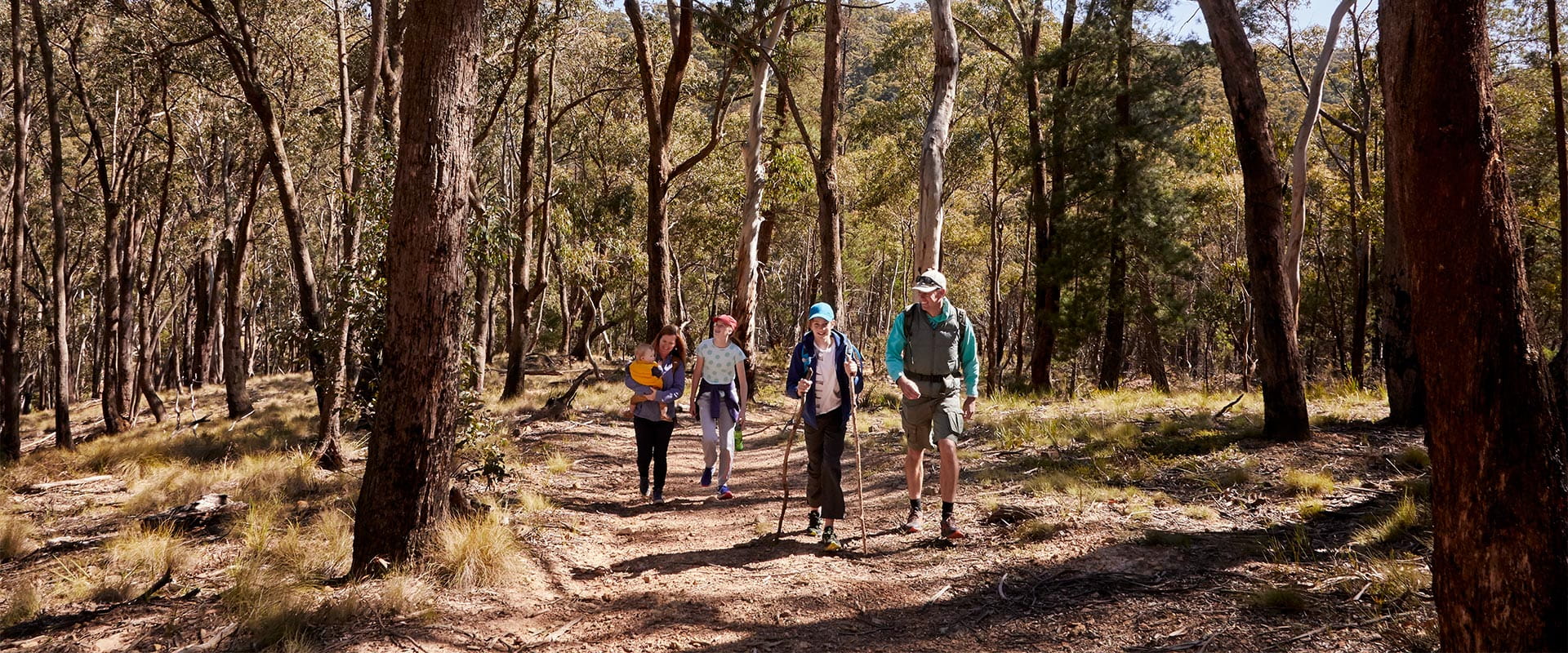 A man wearing hiking clothing smiles at a child in a hat holding two long walking sticks as they walk along a dirt track between trees and vegetation. Following closely behind is a smiling woman holding a baby walking with another child wearing a hat and holding a water bottle.