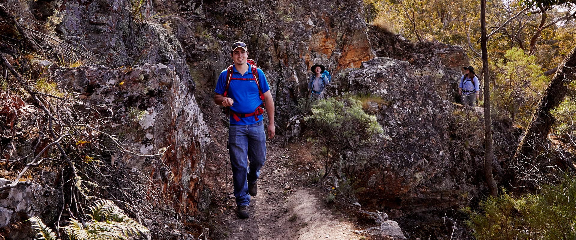 A man in jeans, a short-sleeve blue top, with sunglasses resting on top of a baseball cap, wears a red backpack and holds a hiking stick as he smiles and walks along a dirt track between boulders and vegetation as another two walkers follow behind him.