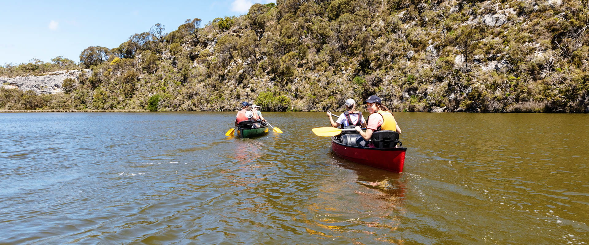 A group of people kayak on the river on a sunny day. One of the kayakers looks back at the camera. 