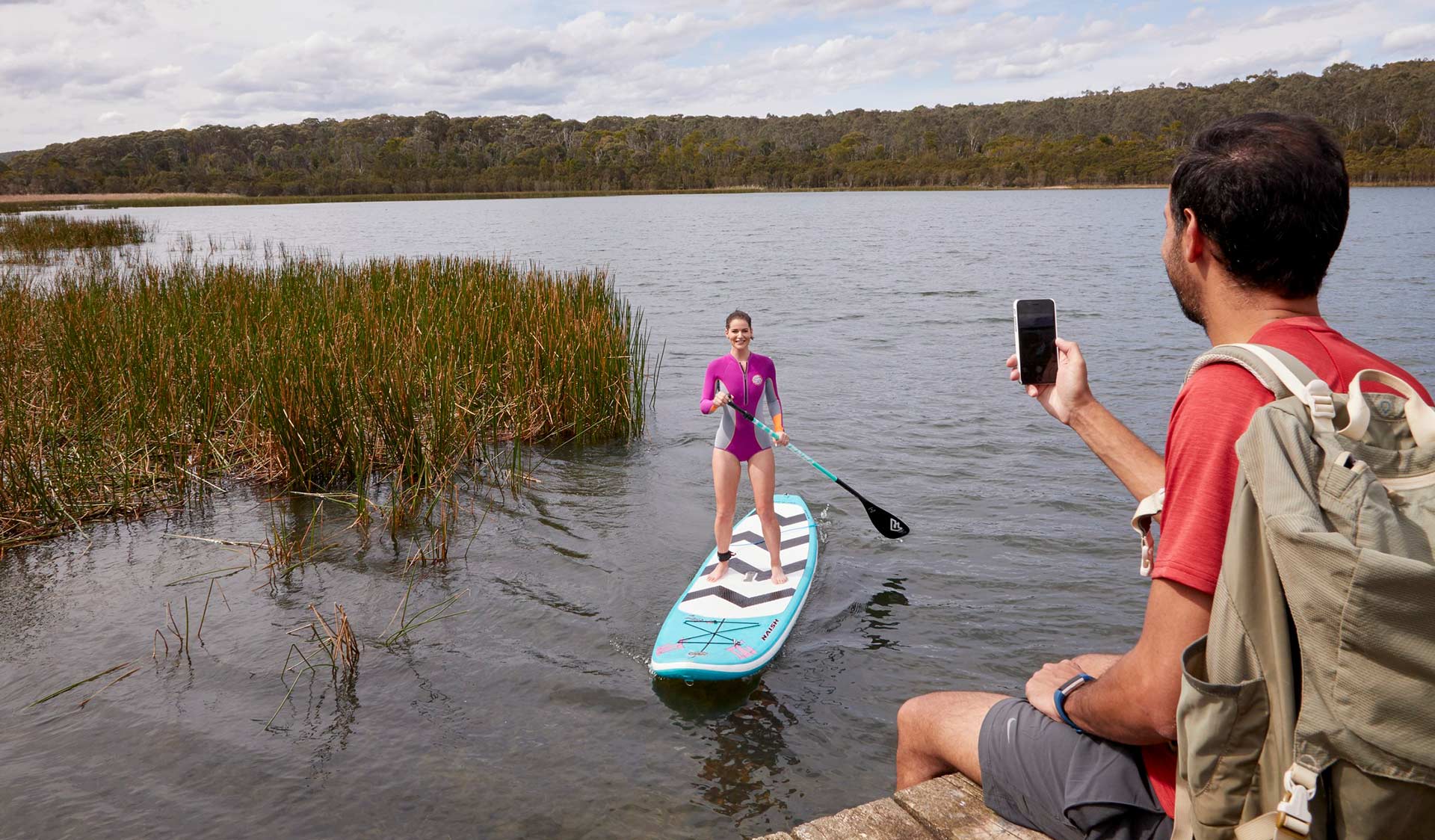 A man takes a photo from the bank of his partner on a stand-up paddle board on Lysterfield Lake.