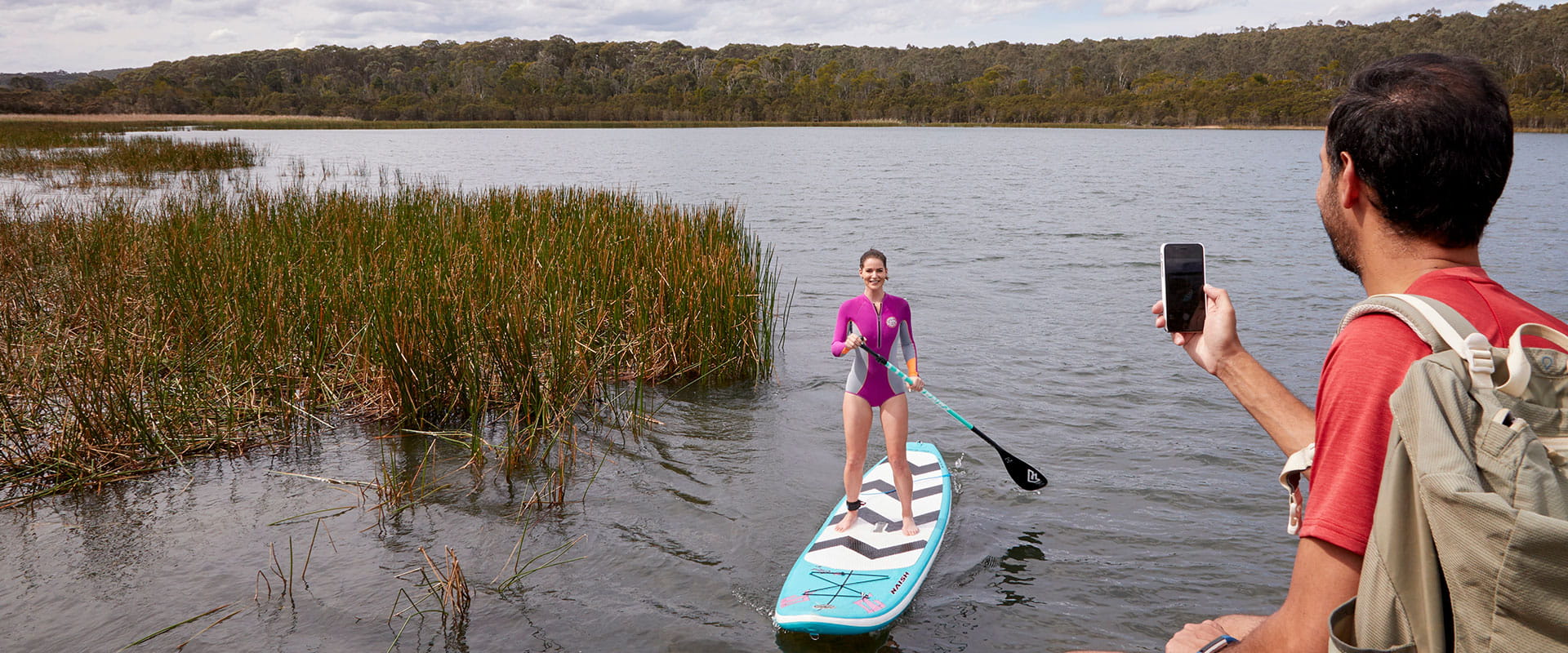 A man takes a photo from the bank of his partner on a stand-up paddle board on Lysterfield Lake.