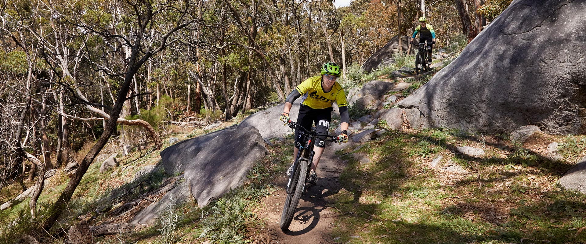 A mountain-biker wearing a helmet and cycling attire rides his mountain bike along a dirt track between boulders, with another cyclist further behind on the trail.