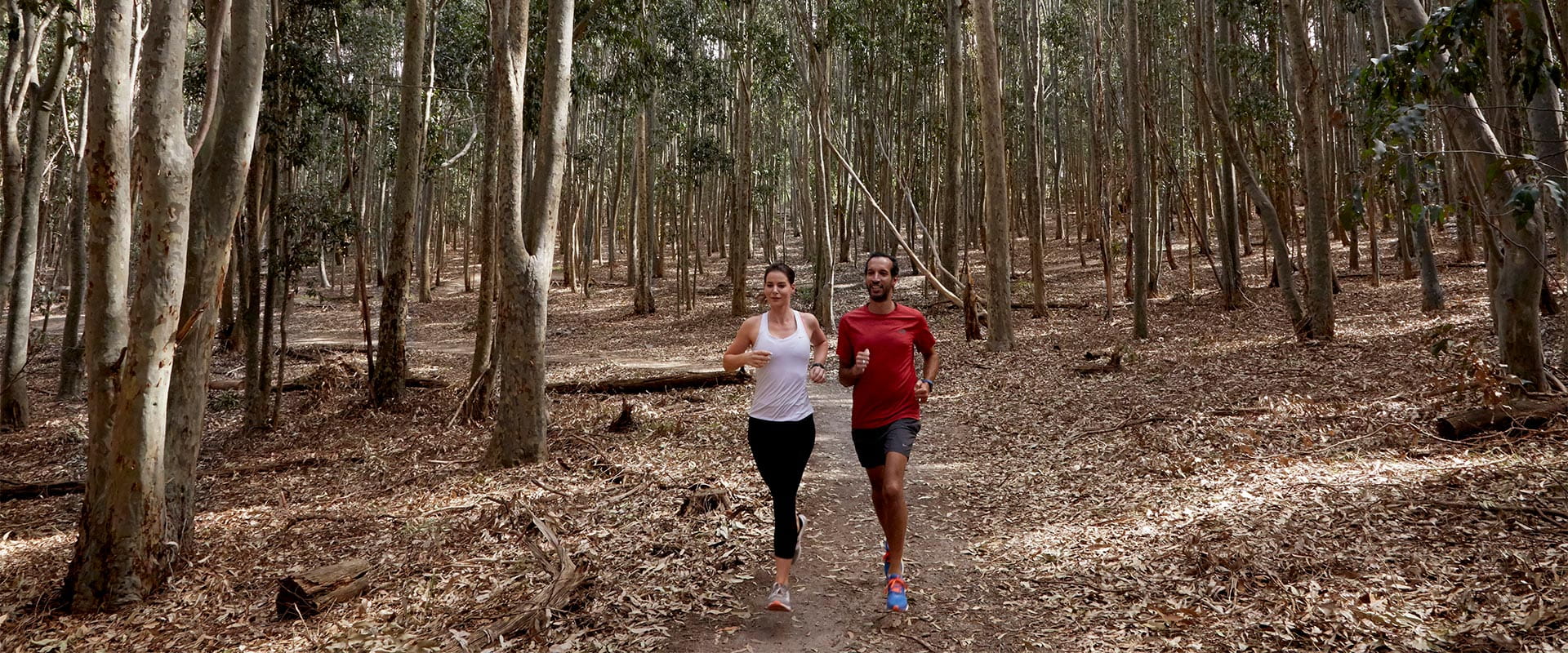A woman and man, both dressed in excercise clothing, run together along a dirt path surrounded by trees.