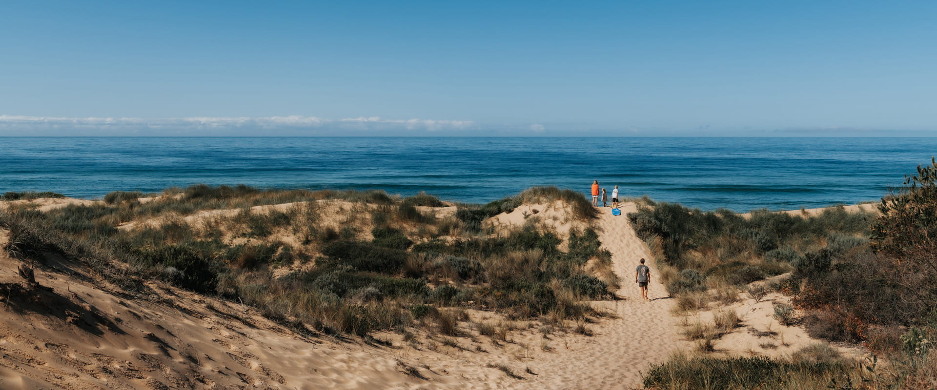 A father walks along a sandy dune path up to his wife and two young children as they stand overlooking the clear blue open waters beyond.