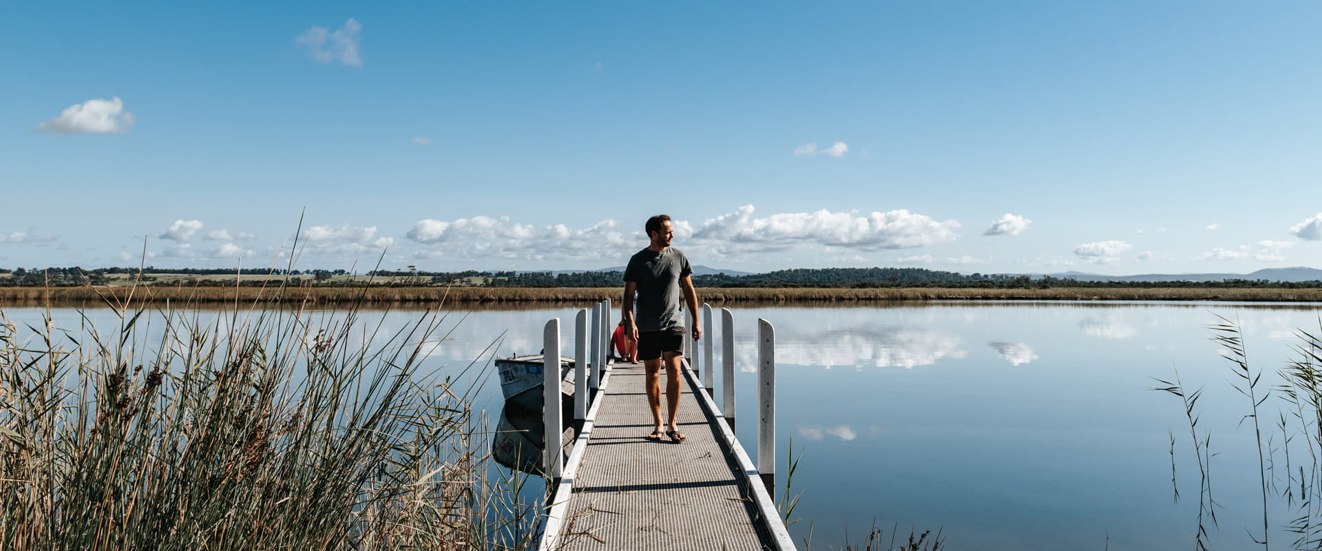 A man walks along a small jetty and looks out over the still waters, with grassland and mountains in the distance behind him.