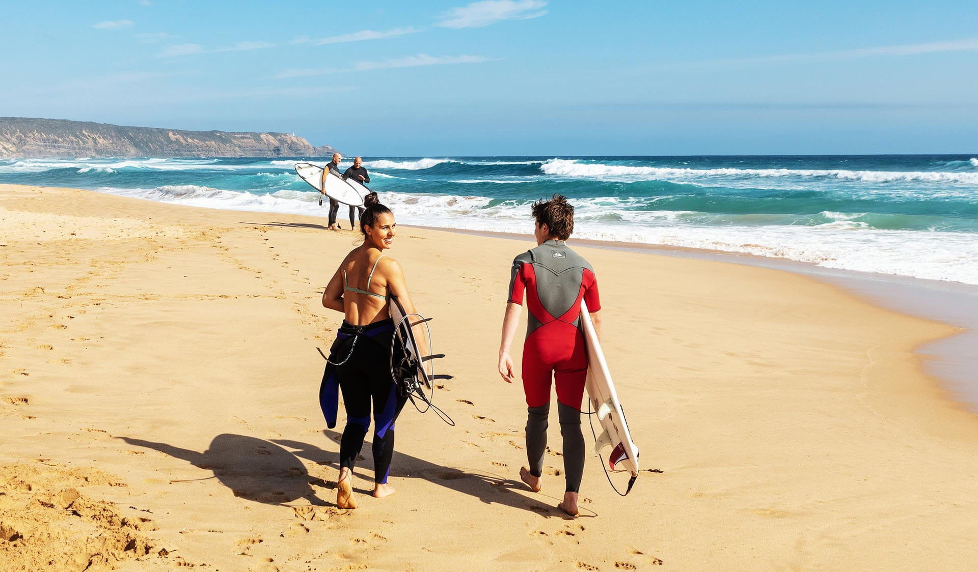 A man in a red wetsuit and a women with the top half of her wetsuit undone follow two men in to the surf on the Morning Peninsula.
