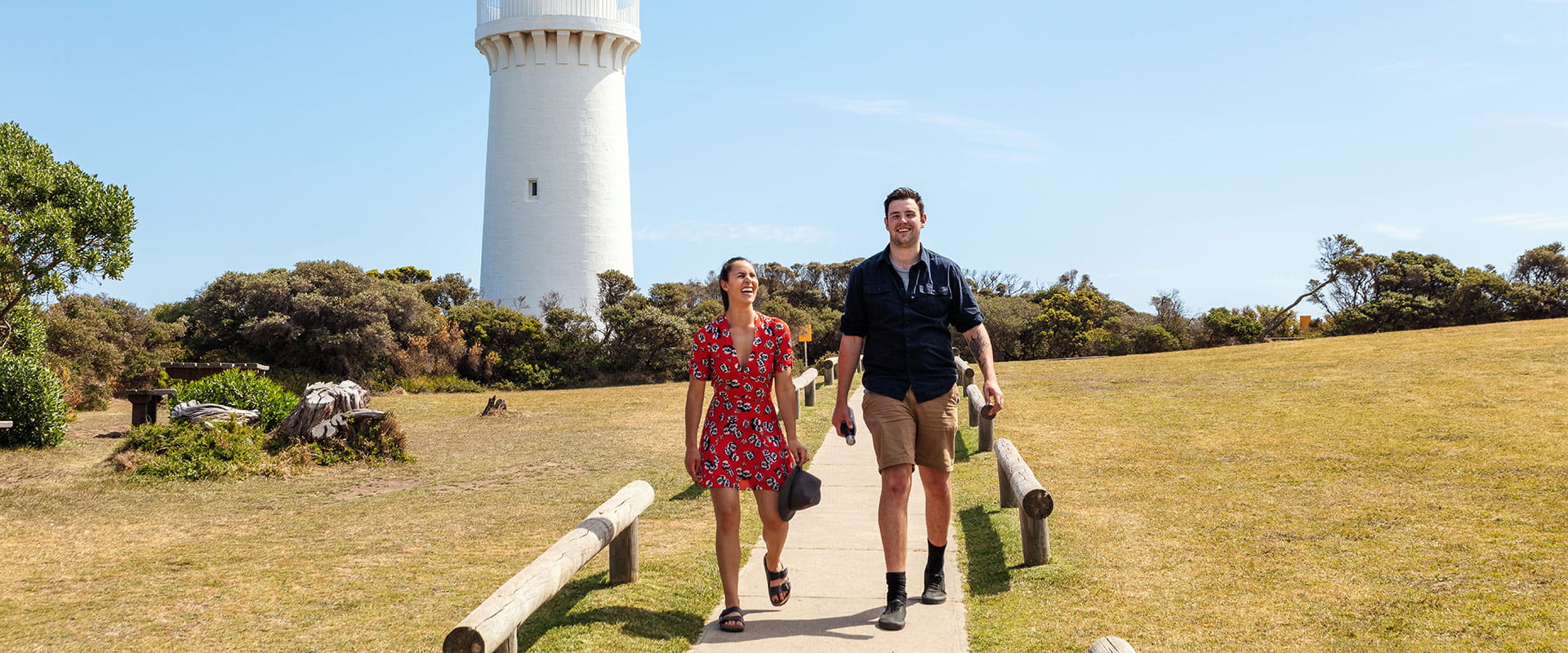 Two people walk away from a large white lighthouse with a red top surrounded by manicured grassland with picnic areas and coastal foliage.