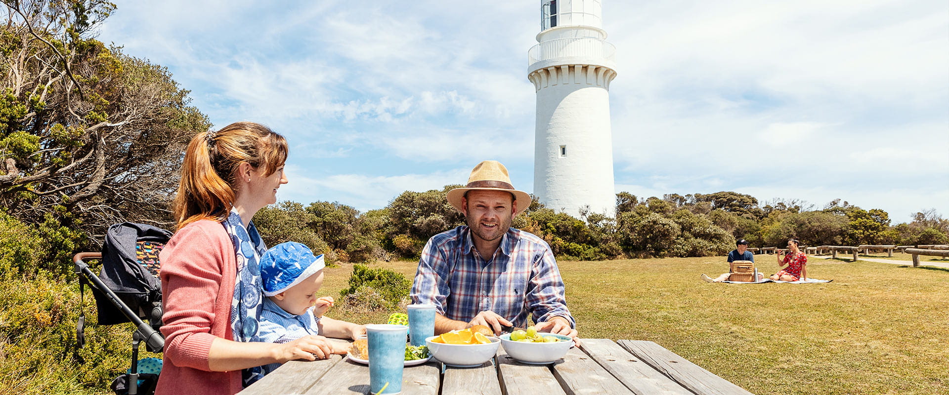 A father, mother and infant child sit at a picnic table infront of a lighthouse