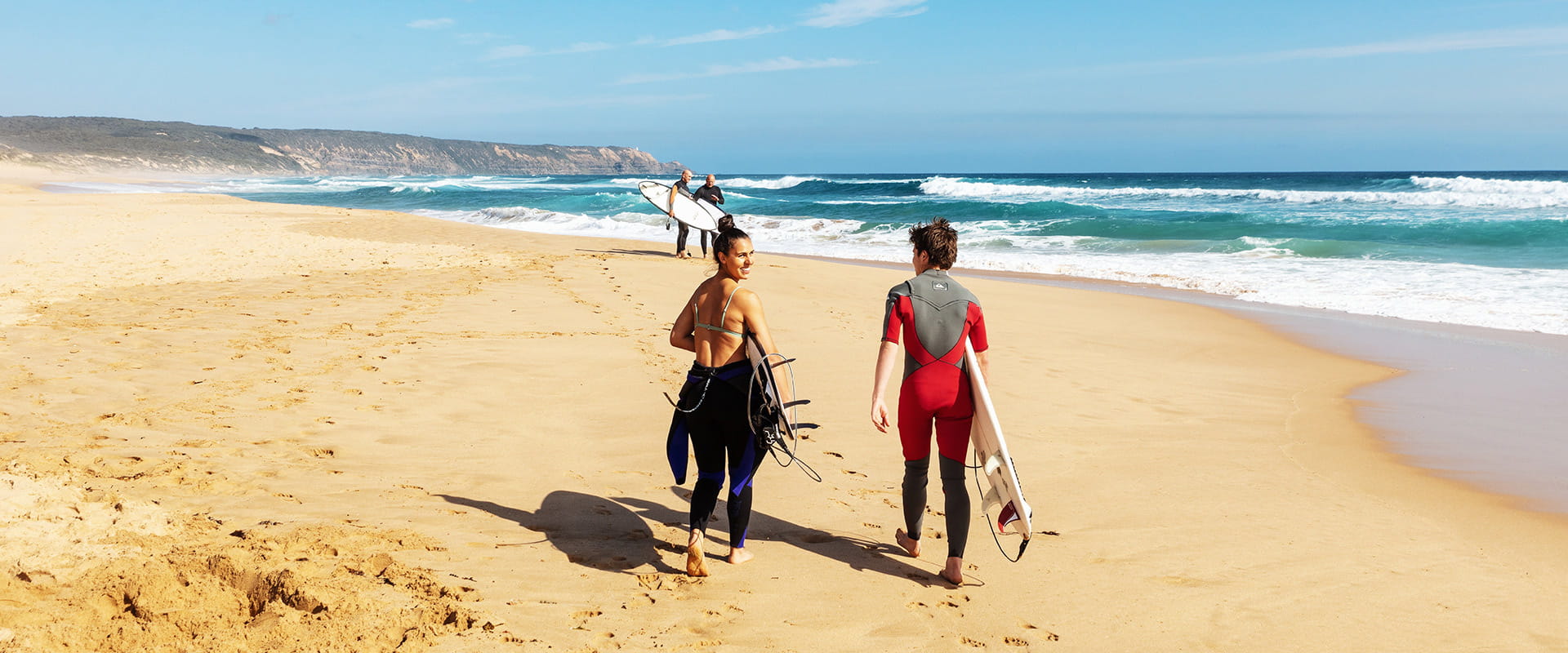Two surfers in wetsuits carring surfboards walk near the waters edge on a sandy surf beach with coastal cliffs in the far distant background.