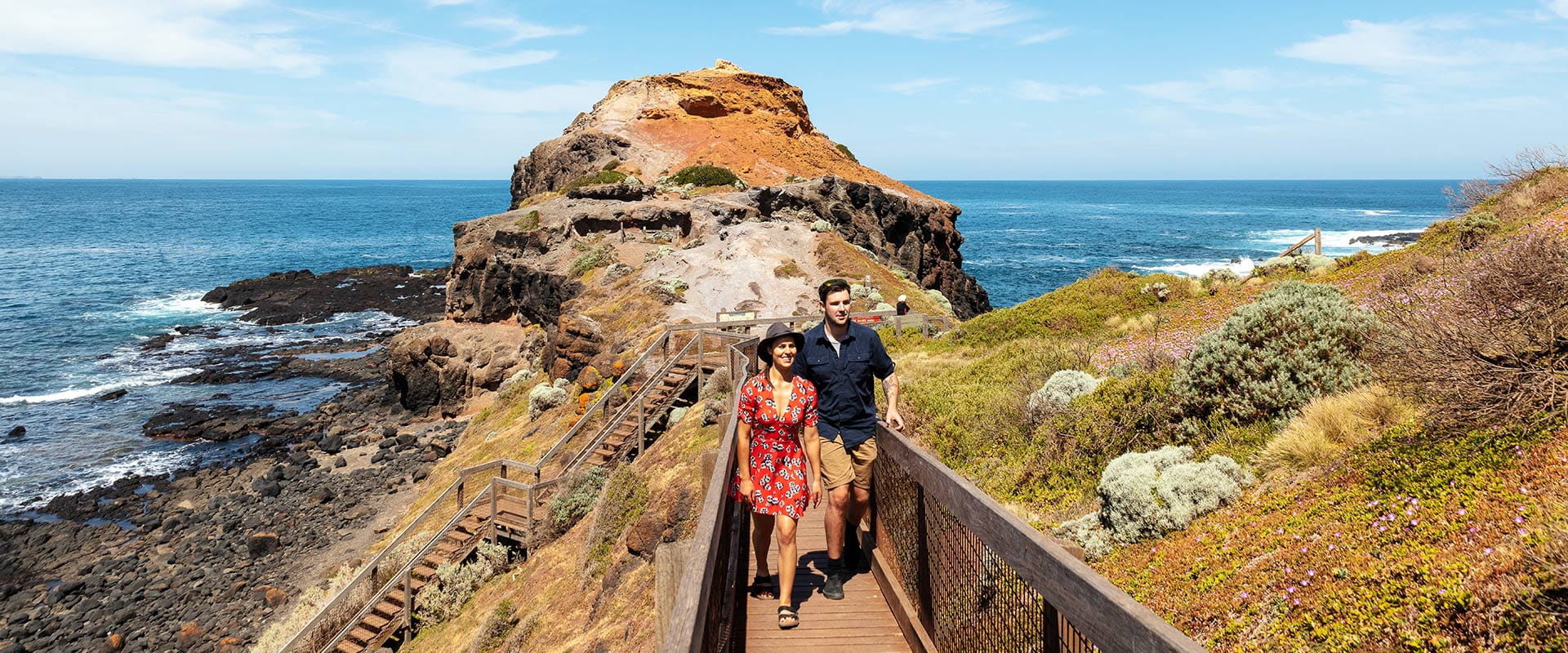 A couple in their twenties walk along a wooden board walk towards the camera in a coastal setting