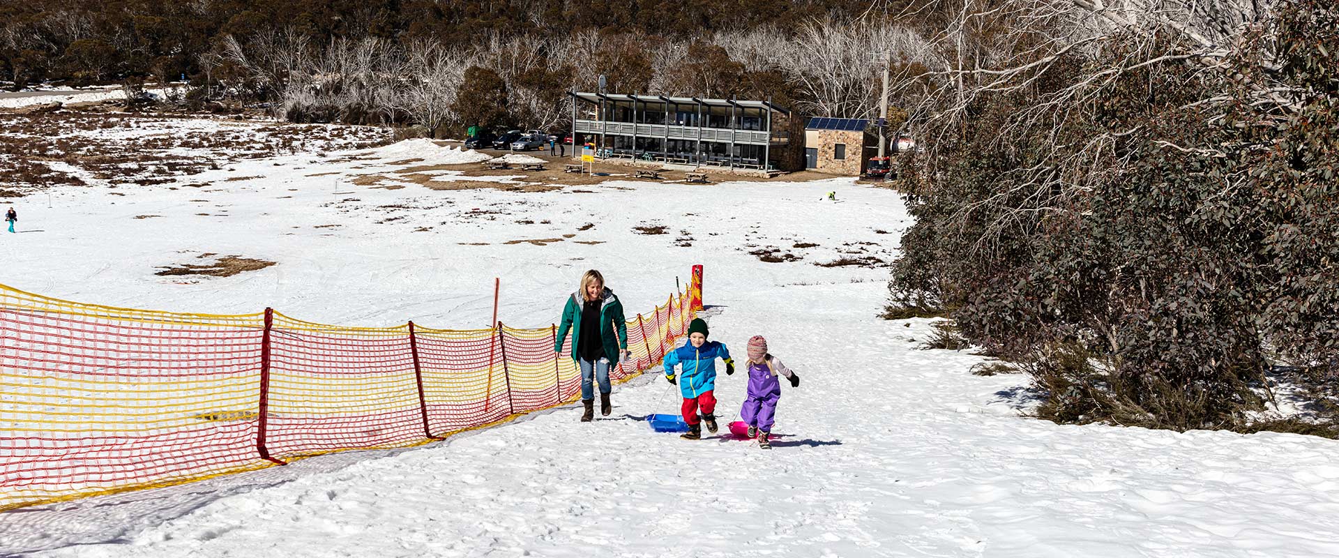 Two young children and their mother head up a gentle snow-cover slope carrying a toboggan