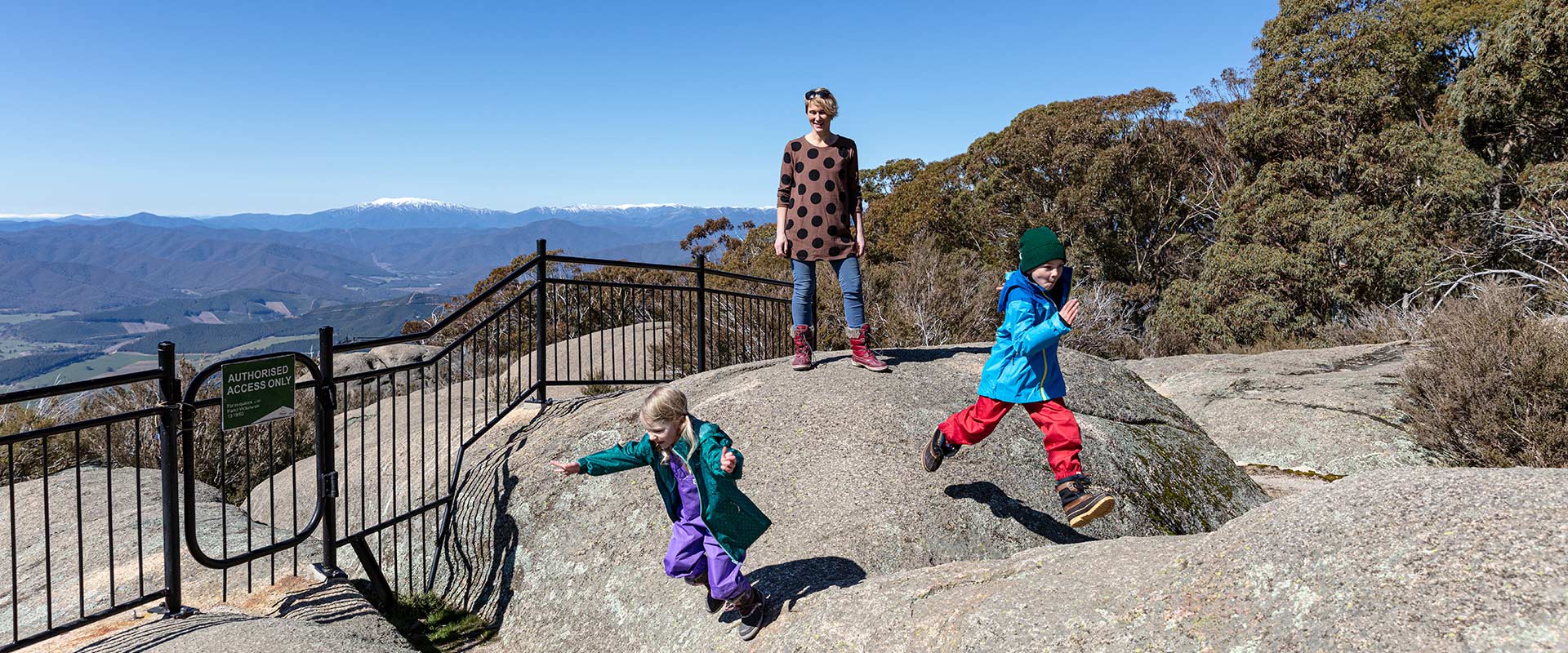 Two small children jump over granite boulders infront of their watching mother, with snow capped mountains in the background